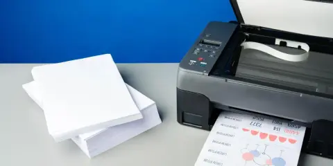 Stos białego papieru obok drukarki, która właśnie drukuje dokument. Idealna klasa papieru do biurowych zastosowań.