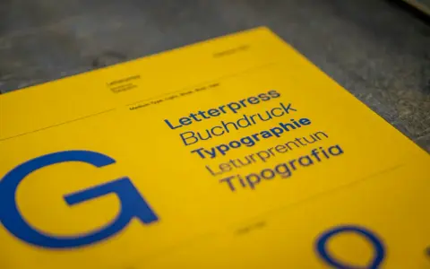 Żółta kartka z niebieskimi napisami: Letterpress, Buchdruck, Typographie, Leturpretun, Tipografia. Duża litera "G" po lewej. To praca typografa.