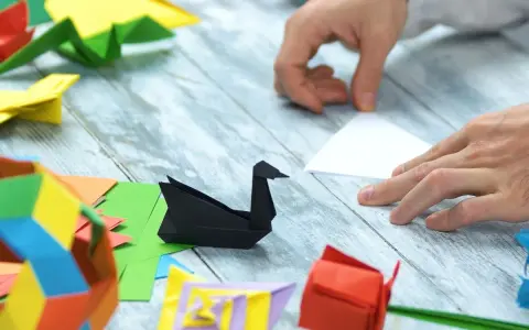 Ręce składają papier, tworząc origami: czarnego łabędzia, czerwoną różę i kolorowe figury. Pokazuje, co można zrobić z papieru.